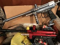 Box containing silver Spyder Xtra and red Spyder Deluxe Compact paintball guns along with various accessories