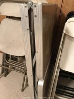 Side view of folded six foot folding table with woodgrain top and metal edging, stacked among folding chairs of plastic and metal.
