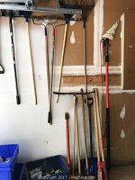 Photo of yard tools hanging and standing against garage wall showing rakes, shovels, post hole digger, and snow shovel.