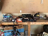 Wide view of long wooden shelf with various scattered tools and hardware items including a drill case, toolbox, and bins filled with miscellaneous items.