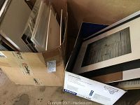 Photo showing multiple framed photographs stacked and leaning in a box and on the floor, some covered in plastic, various sizes.