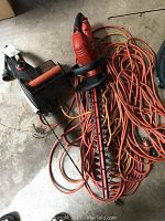 Photo showing the Remington electric chainsaw, Black & Decker hedge trimmer, and multiple orange extension cords tangled on the floor.