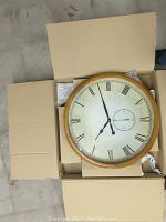 Top view of the large wall clock inside an open box showing the clock face with Roman numerals and secondary seconds dial.