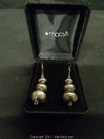 Pair of dangle earrings in black velvet-lined Macy's box. Each earring has three rounded silver segments stacked vertically with engraved floral or sunburst designs.