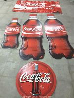 Four Coca Cola plastic advertising banners and one round metal Coca Cola sign laid on floor, showing product details and overall condition.