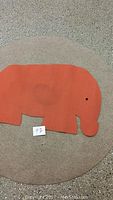 Photo of orange elephant shaped wool felt carpet on top of a round neutral coloured wool carpet on floor.