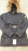 Full view of dark grey Napapijri men's winter coat laid flat showing front pockets, hood with real fur trim, and branded belt buckle with Nordic flag.