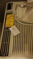 Front view of LG air conditioner showing control panel, front vent, and attached identification tags with model and capacity info.