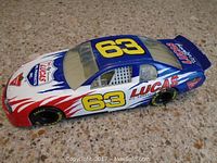 Side view of the die-cast Lucas Oil #63 race car on a speckled surface, showing color and paint details.