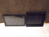 Two black shadow boxes with glass covers placed upright on the floor against a brown backdrop showing front view.
