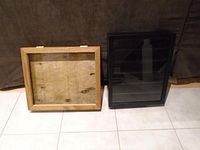 Both shadow boxes side by side on tiled floor against fabric background. Left is natural wood frame, right is black frame.