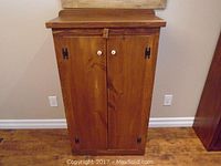 Front view of the pine cabinet showing two doors with white ceramic knobs, locking strap, and black decorative hinges.
