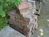 Stacked pile of rectangular bricks showing reddish and gray coloration and wear, beside a concrete slab and greenery