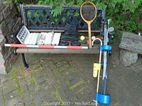Bench with assortment of sporting goods including golf clubs, tennis racket, hockey pucks, plastic golf balls and tees, and a red and white wooden stick.