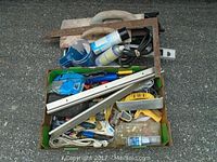 Overall view of box filled with assorted hand tools and accessories including saw, square, gloves, wrench, and more.