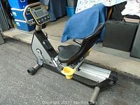 Side view of black and silver recumbent exercise bike with seat, pedals, and digital console visible.