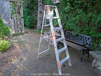 Full view of aluminum step ladder standing open in outdoor setting on brick patio, showing front and side with orange safety markings and wear.