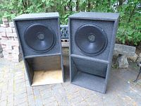 Pair of large black carpeted box speakers with front cone drivers behind black metal grilles and horn-loaded bass ports at bottom. Speakers are outdoors with trees in background.