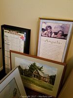 Framed inspirational 'Angels' print with cherubs, rustic house landscape painting, and sports-themed Top 100 Texas plants print leaning against the wall.