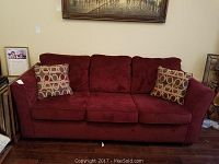 Full frontal image of the burgundy sofa with two throw pillows and framed artwork leaning on wall beside it.
