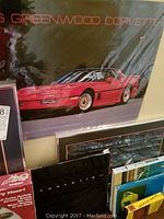 Photo showing a red Corvette poster and some framed prints along a wall with Corvette memorabilia items below