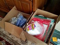 Two cardboard boxes with various paper goods, plasticware, and linens including blue disposable cups, paper doilies, white napkins, and some fabric linens.