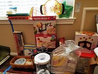 Wide view of kitchen items including apple slicer, Betty Crocker cake decorating box, pans, and other kitchen utensils stacked on a table and floor.