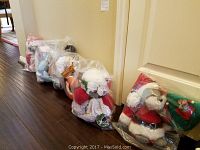 Seven holiday mice dolls individually sealed in clear plastic bags lined up against a wall on a wooden floor.