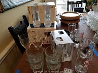 View of glassware on table showing boxed Libbey wine glasses, Anchor Hocking highball glasses, various clear glass mugs, some with Texas A&M logos, and other glassware items.