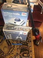 Photo showing Rowenta Commercial Steamer box, Cool Moisture Humidifier box, wrapped accessories, and Eureka vacuum nearby on carpeted floor.