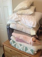 Stack of bedding including pillows, blankets, and folded sheets piled on a wooden dresser showing multiple textures and colors such as white, beige, pink, green and brown