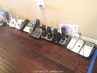 Eleven cordless house phones with charging bases in various styles and brands lined up on wooden floor.
