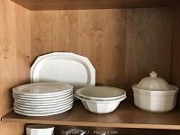 White ceramic Pfaltzgraff dishes including plates, bowls, and tureen arranged on wood shelf.