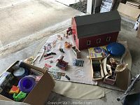 Overall view of red toy barn playset, farm animals, boxed sprinkler, balance cushion, sand toys box, die-cast plane and assorted items on mat