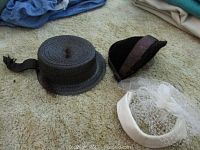 Photo of three vintage women's hats on carpet: one dark woven straw hat, brown bonnet, white hat with netting and flower.