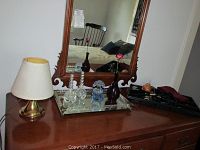 Photo of wooden vanity dresser surface with mirror, showing small brass table lamp, mirror vanity tray with glass perfume bottles and amber vase with flower, and black tray with assorted vintage items including skeleton keys.