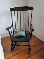 Full view of black wooden rocking chair with gold accents and cushion on seat, placed on wood floor against white wall.