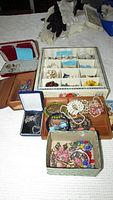 Overview of assorted jewelry including several boxes and trays containing pins, bracelets, earrings, and necklaces, plus a red jewelry case and multiple bracelets and necklaces.