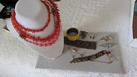 Photo showing coral multi-strand necklace and yellow Bakelite segmented bracelet displayed on white fabric, along with several decorative pins and brooches.