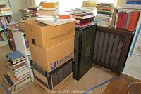 Various hardcover and softcover books stacked and shelving, with boxes and pile of books beside them.