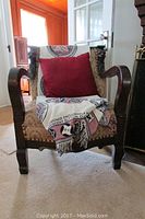 Front view of vintage carved wood framed upholstered chair with a red pillow and decorative blanket placed on it, showing carved wood armrests and fabric upholstery.