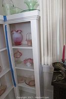 Shelves displaying various vintage glass pieces including pink hobnail glass vases and bowls with ruffled edges, and green glass items.