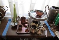 Wide view of sterling silver and mixed metal tableware and candle holders on a wooden surface with two green candles in candle sticks, sugar bowl, creamer, pencil cases, candy dish, candle snuffer, and tray.