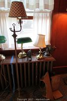 Three table lamps on radiator: tall metal lamp with brown patterned shade, brass banker's lamp with green glass shade, small brass lamp with white shade and wire fan behind