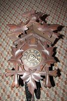 Front view of the wooden cuckoo clock showing the carved leaves, clock face with Roman numerals, and the top with a bird figure.