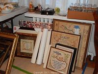 Needlework pieces and rolled textiles leaning against a wall showing two framed vintage needlework pieces with embroidery and framed posters behind.