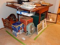 Wide shot of the lot showing a wooden table holding various linens, boxes and framed artworks on the floor including a boxed Oster blender and Mirro percolator box, a blue vintage fan, and assorted framed pieces leaning against the table and wall.