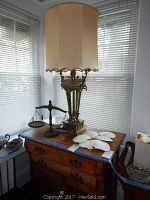 Ornate brass base table lamp with beige fabric shade on wooden dresser next to wooden pan balance scale and several white scallop shells.