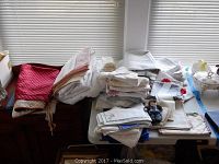 Wide view of folded table linens, napkins, and cloths stacked on table beside window. Mixture of white and colored fabrics.