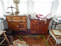 Photo showing vintage wooden wheeled chest with two drawers and doors next to a wooden wash stand with drawer and doors, both topped with various items and textiles.